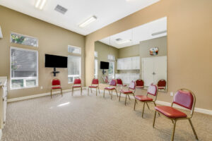 Interior Community Meeting room, large mirrored wall, contemporary carpeting, neutral toned walls, tv on wall, 7 red chairs in a circle.
